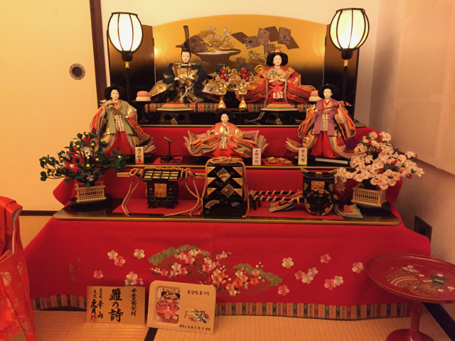 The Hina dolls: a princess and prince on the top shelf, with three female attendants on the shelf below, and an ox-drawn carriage, palanquin, and treasure box on the bottom shelf. All the dolls are dressed for the Heian period.