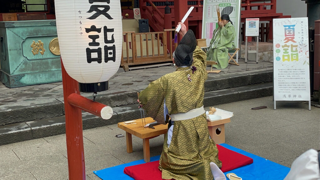A man in Heian-period Japanese dress is kneeling in front of a low wooden table, set in front of the prayer hall of a jinja. An eel is visible on the table, and the man is waving a long knife over his head.